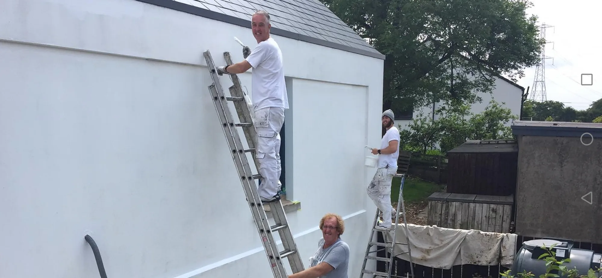 Three of the team painting the white wall of a building from ladders, smiling at the camera